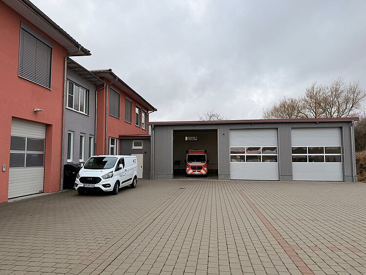 Rettungswache Blaufelden Außenansicht der Rettungswache in Blaufelden. Man sieht drei Garagen, eine davon ist offen. Links von den Garagen ist die ein rotes zweistöckiges Gebäude zu sehen - die Rettungswache.